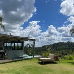 Área gramada com espreguiçadeira, casa ao lado, piscina parcialmente visível e céu com nuvens.